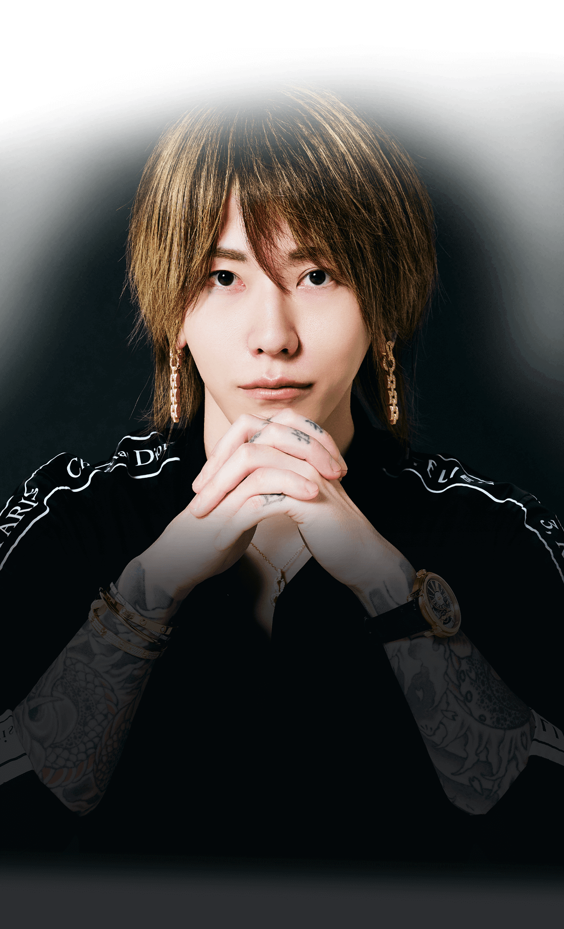 黒い背景で腕を組み見つめるおじゃる丸社長。ファッション性の高いタトゥーとアクセサリーが印象的なポートレート写真。