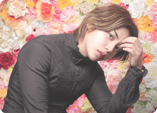 花を背景に黒い服を着た一ノ瀬 寅がポーズをとるポートレート写真。華やかな雰囲気とエレガントな印象を与えるプロフィールイメージ。